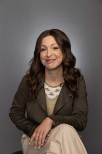 A woman with long brown hair sits on a stool against a gray background, wearing a brown jacket, beige dress, and a statement necklace, smiling softly with her hands folded.