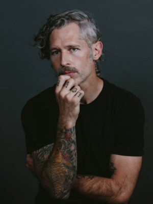 David Walter Banks A man with gray curly hair, a mustache, tattoos on his arms, and rings on his fingers, wearing a black shirt, poses with his hand on his chin against a dark background.