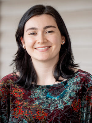 Erin Cribbs, a woman with straight dark hair, wearing a floral-patterned top, stands outdoors in front of a neutral-colored wall and smiles at the camera.