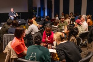 A group of people sit at tables in a dimly lit room, talking and facing a person speaking at a microphone on stage.