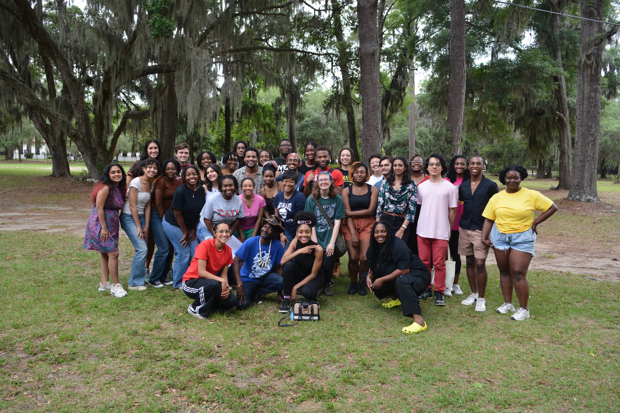 A large group of people poses together outdoors on grass with trees in the background, some standing and some kneeling, smiling at the camera.