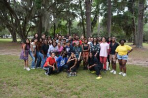 A large group of people of diverse ages and backgrounds poses together outdoors in a wooded park area, smiling at the camera.