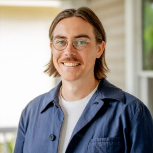 A person with glasses and a mustache smiles at the camera, wearing a white shirt and a dark blue jacket, standing in front of a blurred outdoor background.