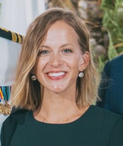 A woman with shoulder-length blonde hair, wearing earrings and a dark green top, smiles at the camera in an outdoor setting.