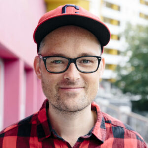 A person wearing glasses, a red plaid shirt, and a red cap stands outdoors in front of a pink wall and a blurred building background.