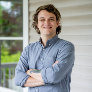 A person with wavy brown hair, wearing a light blue button-up shirt, stands outside in front of a house siding and window, smiling with arms crossed.