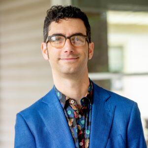 A man with short curly hair and glasses wears a blue suit jacket and a patterned shirt, standing outdoors in front of a blurred background.