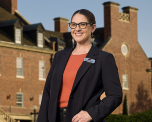 A woman wearing glasses, a black blazer, and an orange shirt stands smiling in front of a large brick building on a sunny day.