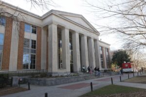 A large neoclassical building with tall columns at the entrance; several people walk up the steps, and a red truck is parked nearby.
