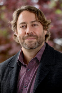 A man with medium-length brown hair and a beard wearing a dark blazer and patterned shirt stands outdoors in front of blurred foliage.
