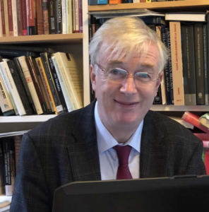 A man with gray hair and glasses, wearing a dark jacket and red tie, sits in front of a laptop with bookshelves in the background.