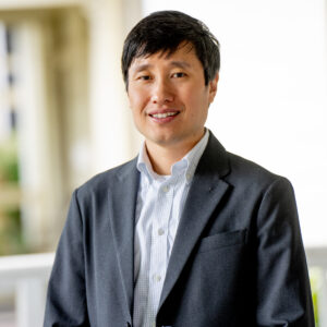 A man in a dark suit jacket and light dress shirt stands indoors, smiling at the camera with a blurred background.