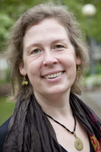 A woman with wavy light brown hair smiles outdoors, wearing gold earrings, a dark scarf, and a spiral pendant necklace.