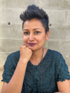 A person with short, spiked hair and dangling earrings sits at a table, resting their chin on their hand, in front of a concrete wall.