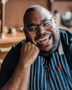 A man wearing glasses and a striped apron smiles broadly while resting his chin on his hand, seated in a warmly lit room.