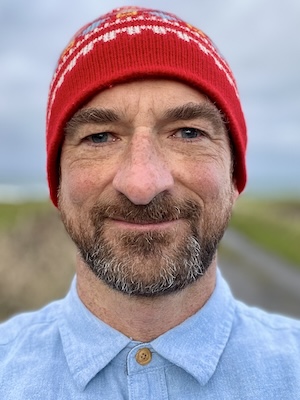 A man with a beard wearing a red knit beanie and a light blue button-up shirt is standing outdoors, smiling at the camera.