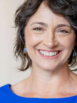A woman with short, wavy dark hair and blue earrings smiles at the camera, wearing a sleeveless blue top against a plain background.