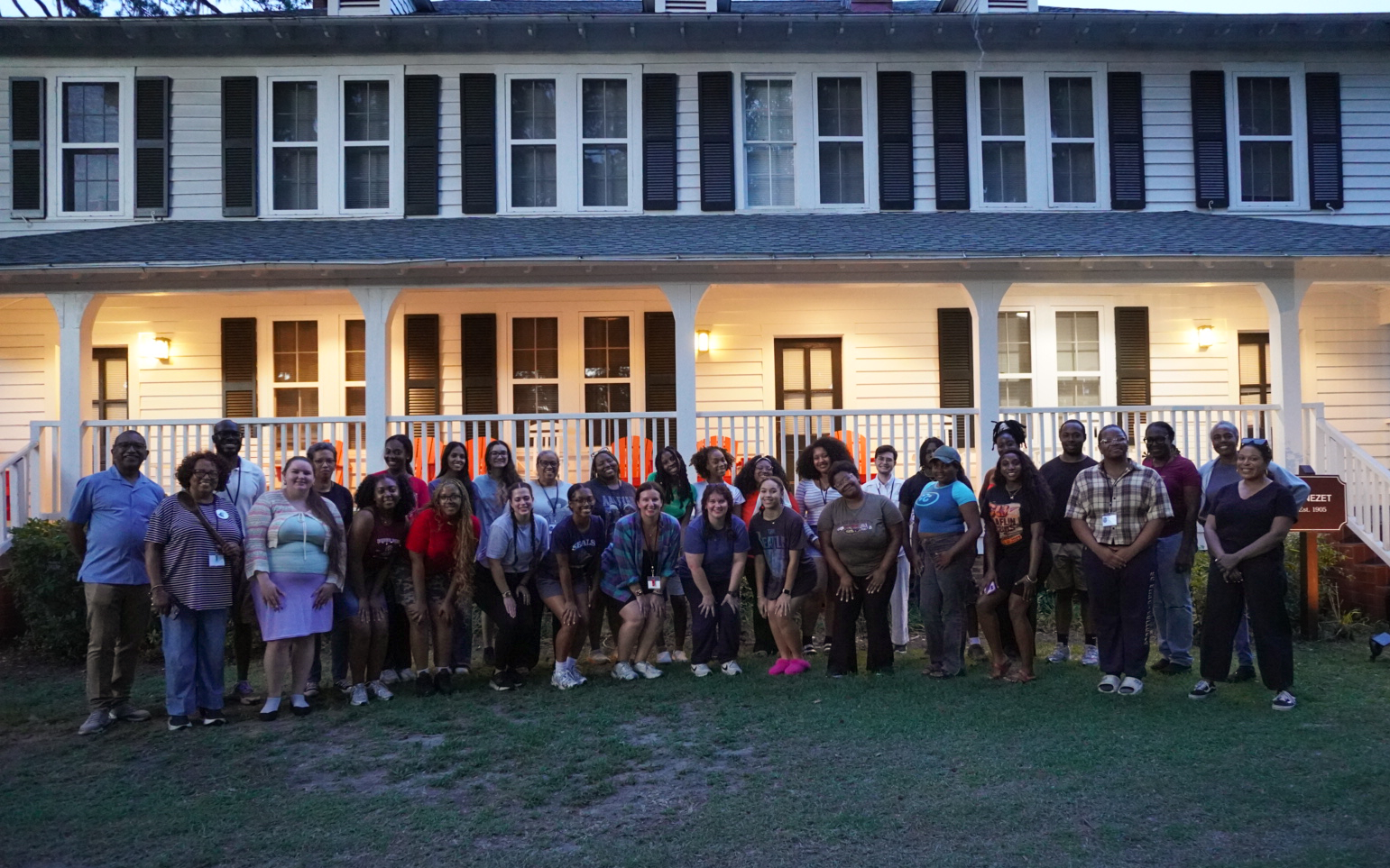 A large group of people poses for a photo in front of a two-story house with white siding and black shutters at dusk.