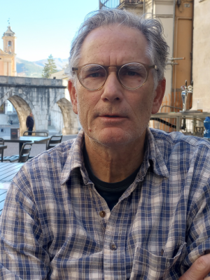 An older man with glasses and a plaid shirt sits at an outdoor café table; historic buildings and an arched stone structure are in the background.