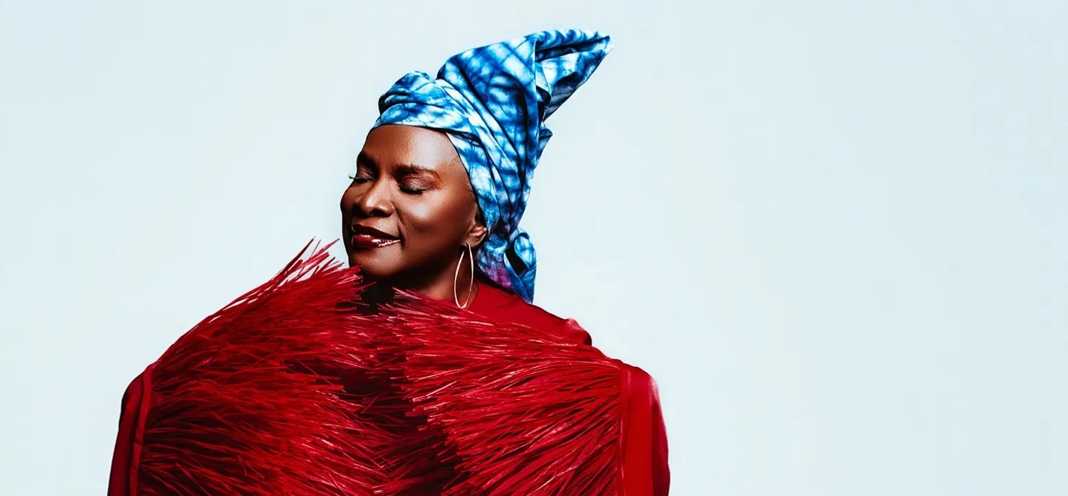 A person in a red textured outfit and blue patterned headwrap stands against a plain light background, eyes closed and smiling softly.