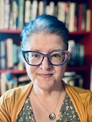 Elise Robinson Person with short blue hair, glasses, and a nose ring, wearing a yellow cardigan and floral dress, smiles in front of a blurred bookshelf background.