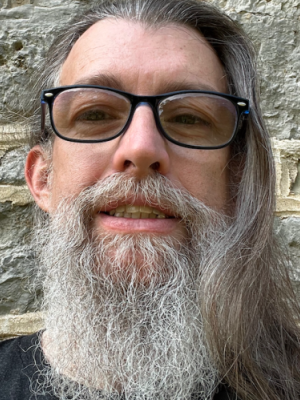 James Owen A man with long gray hair, a full gray beard, and glasses stands in front of a stone wall, looking at the camera.
