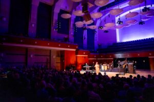 Musicians perform on a brightly lit stage in a large concert hall with a seated audience and purple lighting above.