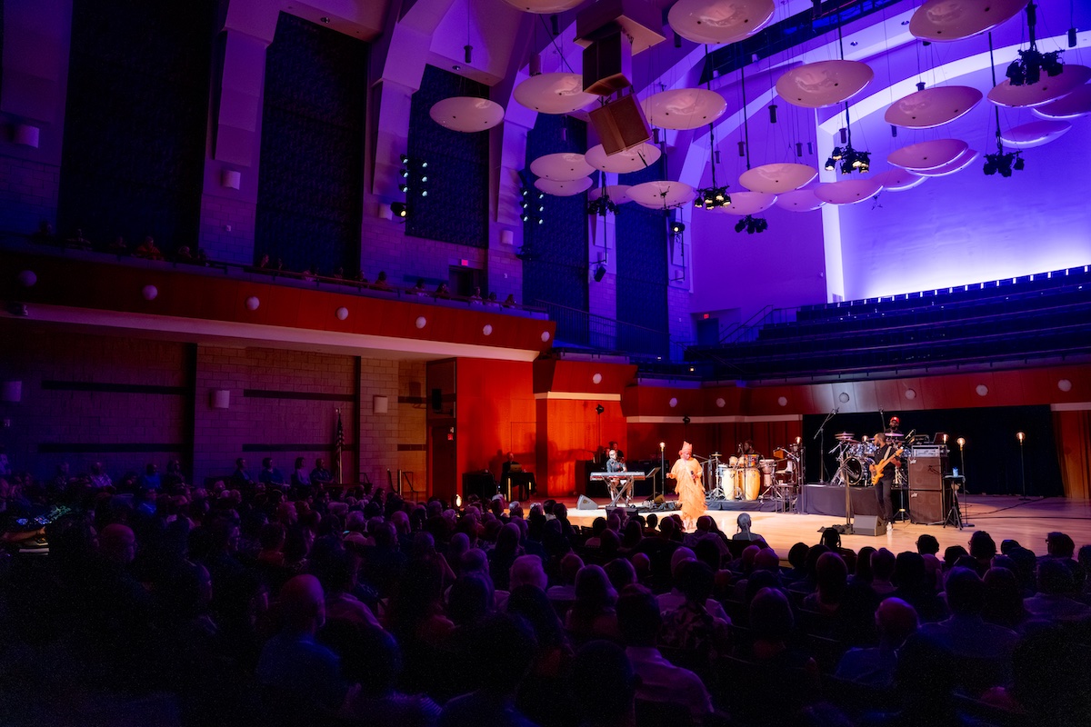 Musicians perform on a brightly lit stage in a large concert hall with a seated audience and purple lighting above.