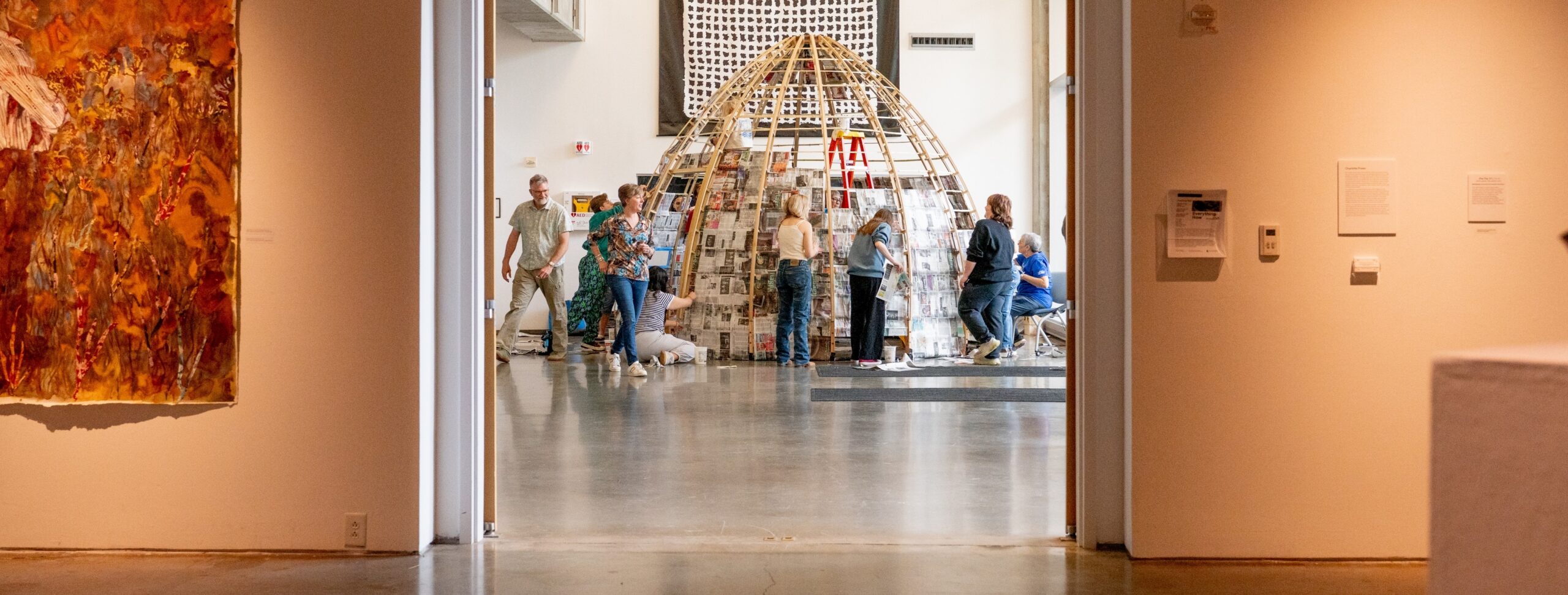 People gather around a dome-shaped structure covered in newspapers inside an art gallery with white walls and artworks on display.