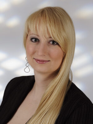 Franziska Naether A woman with long blonde hair and side-swept bangs, wearing a black blazer and drop earrings, poses in front of a softly lit background.