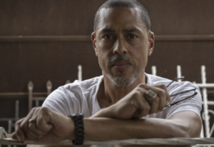A man with short hair and a goatee leans on a railing, holding eyeglasses in one hand and wearing a white shirt, ring, and watch.
