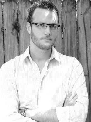 Man with short hair and glasses stands in front of a wooden fence, wearing a striped shirt with arms crossed, looking at the camera.