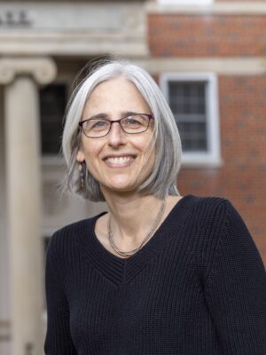 Rachel Gabara A woman with gray hair and glasses stands outdoors in front of a brick building with columns, wearing a black sweater and smiling at the camera.