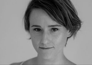 A person with short, tousled hair looks directly at the camera with a neutral expression. The photo is in black and white.