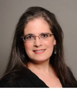 Woman with long dark hair and glasses, wearing a black top and earrings, smiling against a plain gray background.