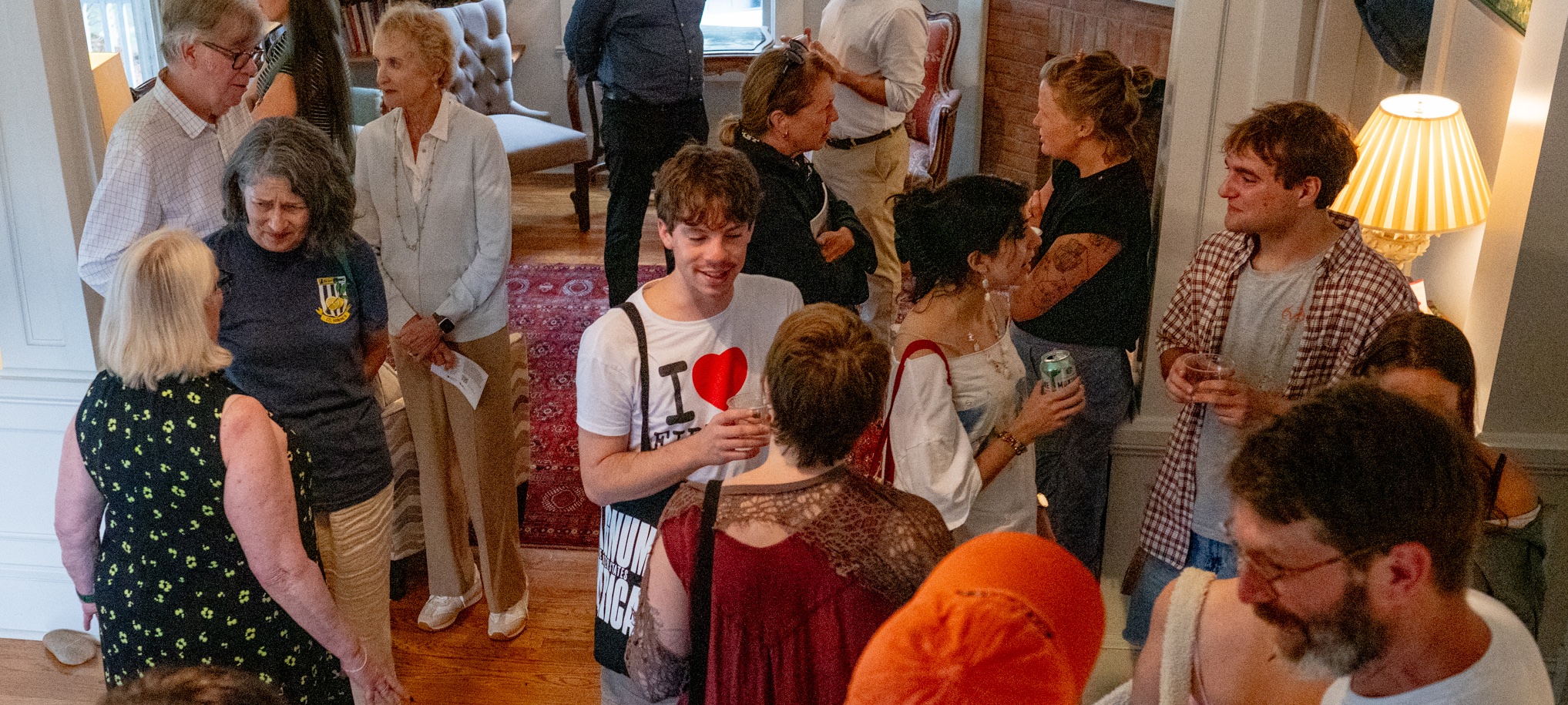 A group of people socialize and converse in a warmly lit living room, some holding drinks, while standing and mingling in small groups.