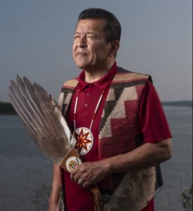 Dwayne Tomah An Indigenous man in a red polo and patterned vest holds a ceremonial fan of feathers.