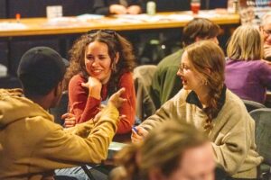 Humanities Trivia GOOD Three people sitting at a table together in a dimly lit room, two women smiling while talking to a man in a tan hoodie.
