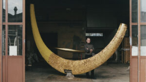 Joseph Walsh A man stands with arms crossed behind a large, gold, crescent-shaped sculpture inside a workshop.